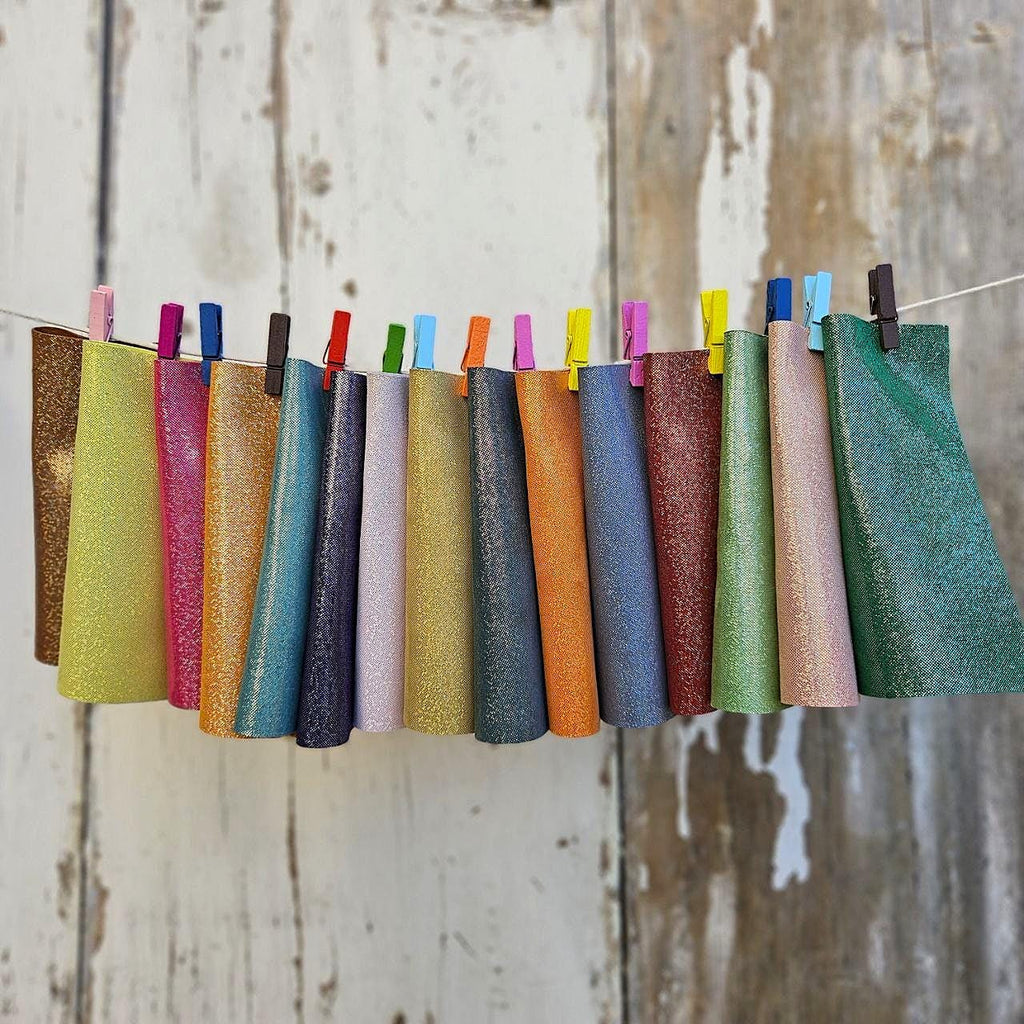 Può includere: Una collezione di coni di tessuto glitterato colorati appesi a un filo con mollette colorate. I coni presentano una gamma di colori, tra cui verde, rosa, blu e arancione, su uno sfondo di legno bianco invecchiato.