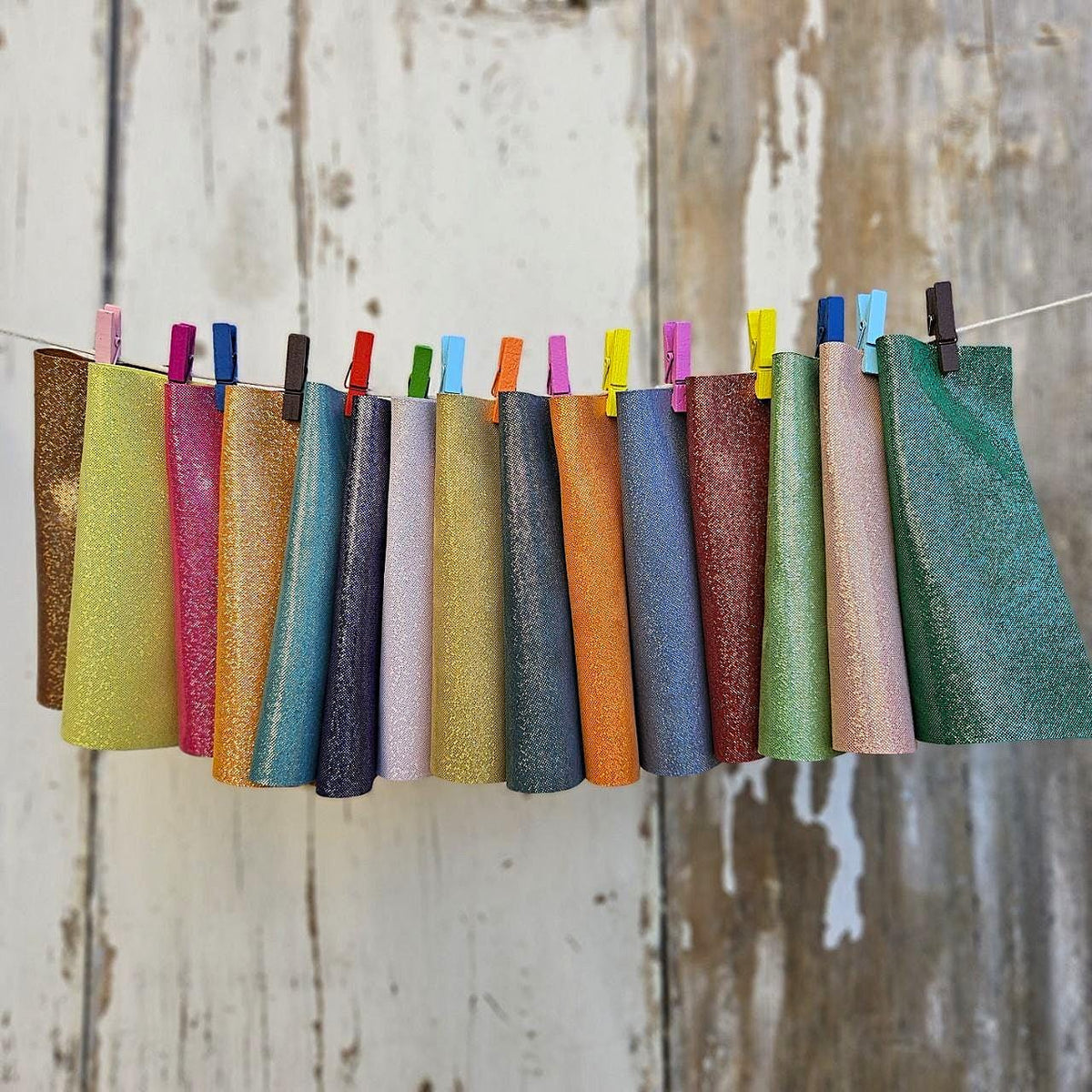 Può includere: Una collezione di coni di tessuto glitterato colorati appesi a un filo con mollette colorate. I coni presentano una gamma di colori, tra cui verde, rosa, blu e arancione, su uno sfondo di legno bianco invecchiato.