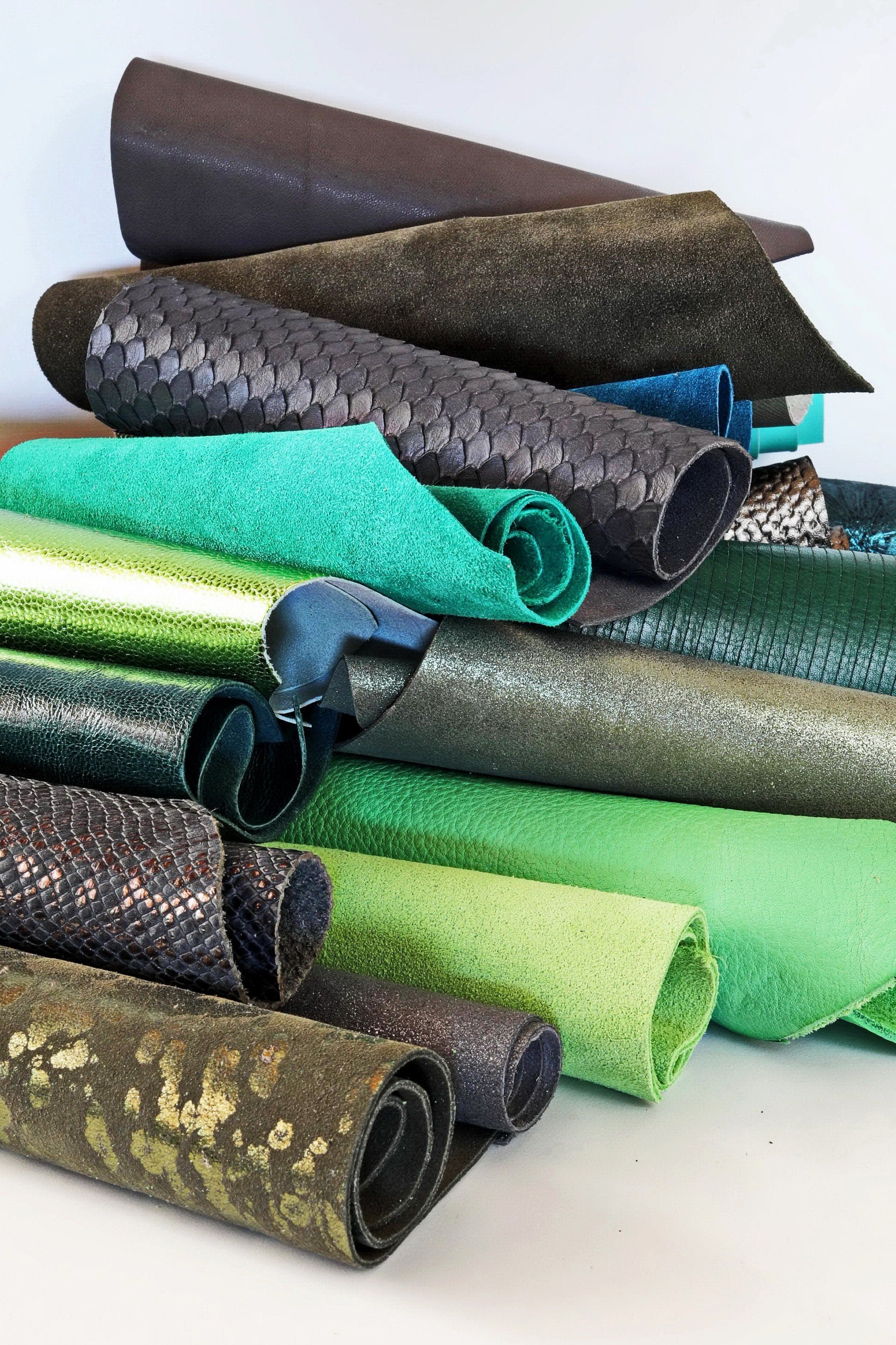 Può includere: Una collezione di pelle arrotolata in vari colori e trame, tra cui verde, marrone, nero e grigio. La pelle è disposta in una pila, con i rotoli impilati uno sopra l'altro.