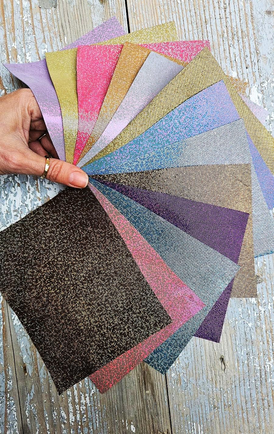 Può includere: Una mano tiene un ventaglio di campioni di tessuto glitterato in vari colori. I campioni includono tonalità di rosa, oro, argento, blu e viola. I tessuti hanno un aspetto strutturato e scintillante, disposti su una superficie di legno invecchiato.