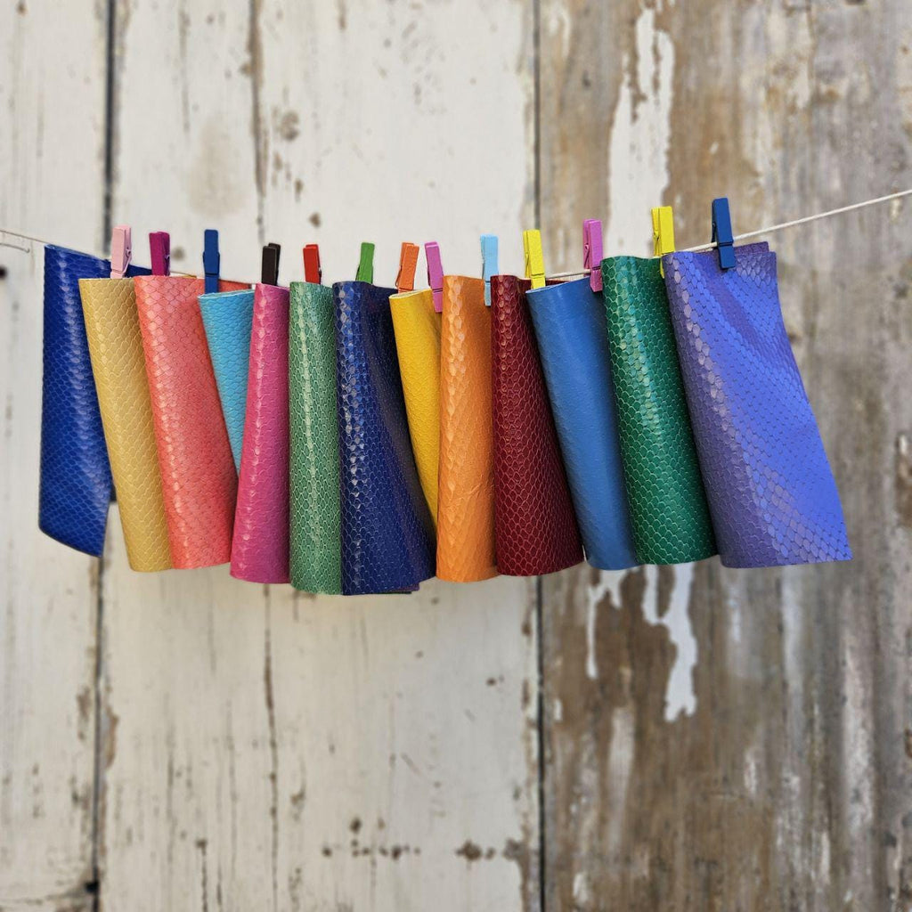 Può includere: Una collezione di rotoli di tessuto colorati, ciascuno fissato con una molletta e appeso a uno spago. I tessuti mostrano una varietà di colori vivaci, tra cui blu, giallo, arancione, rosso e verde, su uno sfondo di legno rustico e invecchiato.