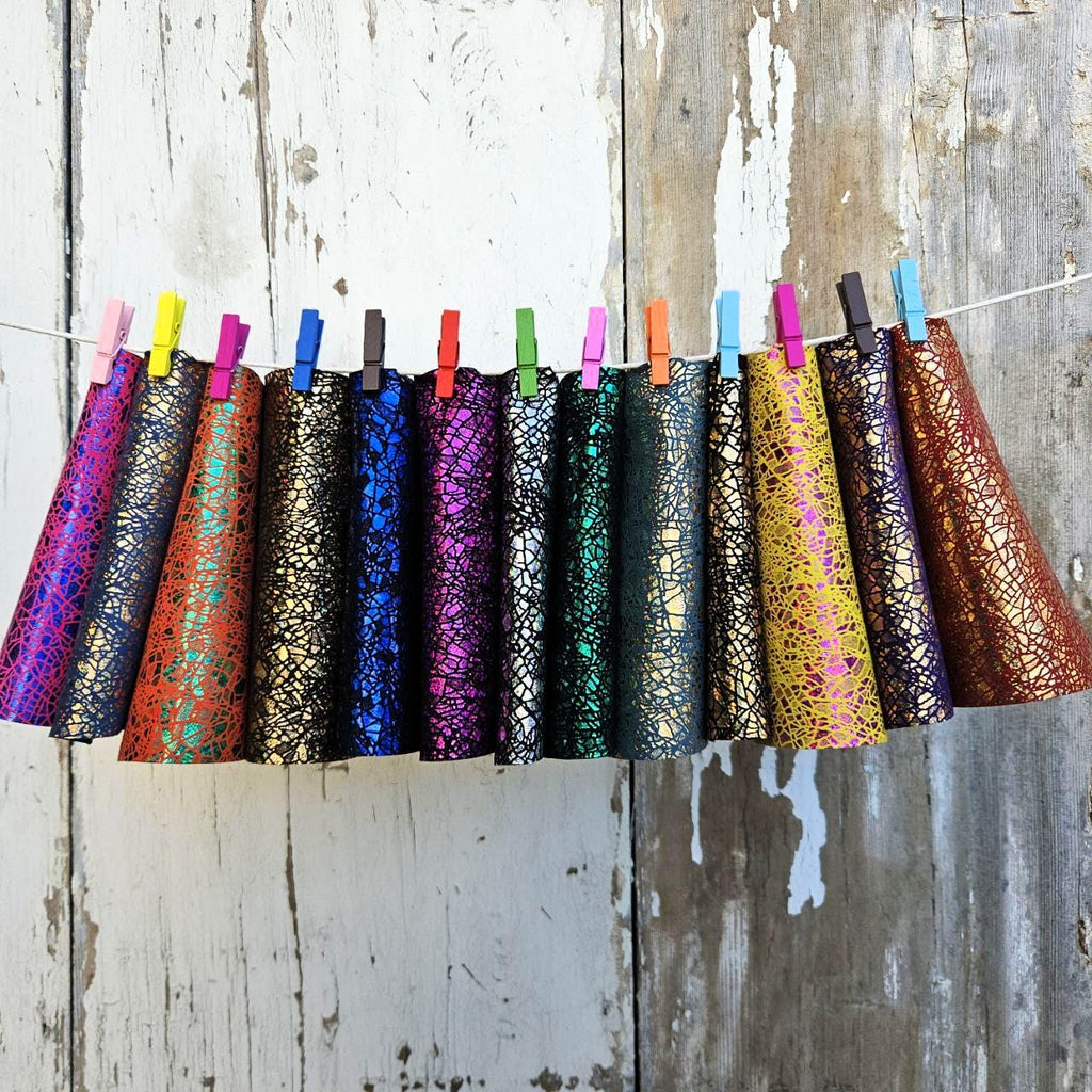 Può includere: Una fila di cappelli da festa a forma di cono colorati appesi a una corda con mollette. I cappelli sono in vari colori, tra cui viola, rosso, blu, verde e oro, con una finitura metallica strutturata e screpolata. Lo sfondo è una parete di legno bianco invecchiato.