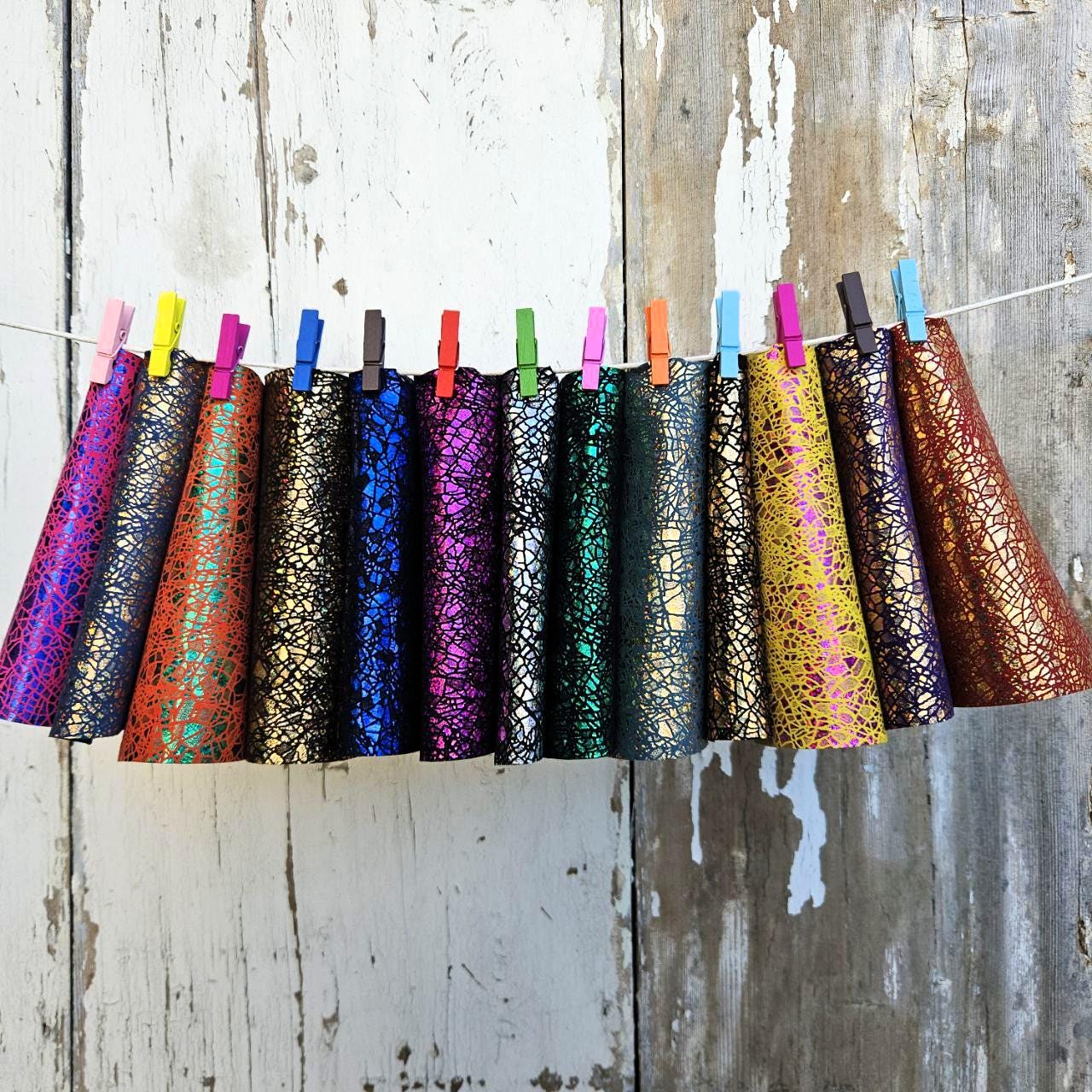 Può includere: Una fila di cappelli da festa a forma di cono colorati appesi a una corda con mollette. I cappelli sono in vari colori, tra cui viola, rosso, blu, verde e oro, con una finitura metallica strutturata e screpolata. Lo sfondo è una parete di legno bianco invecchiato.