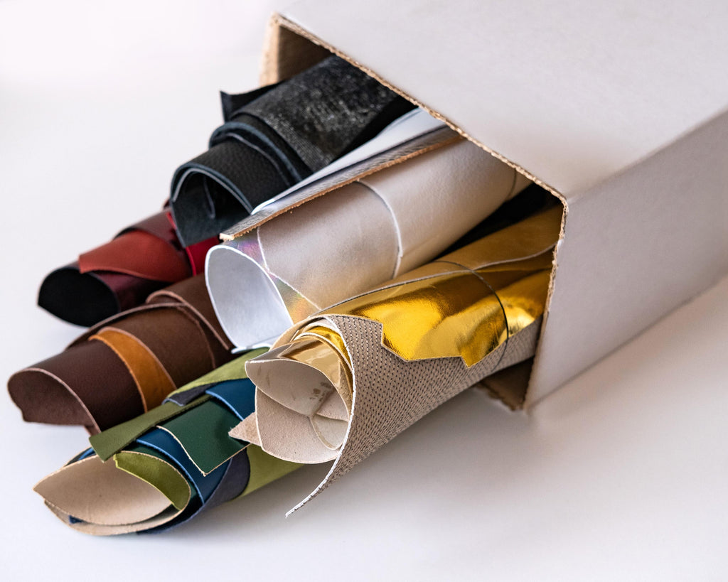 Può includere: Una scatola piena di rotoli di tessuti di diversi colori, tra cui marrone, oro, bianco e verde.