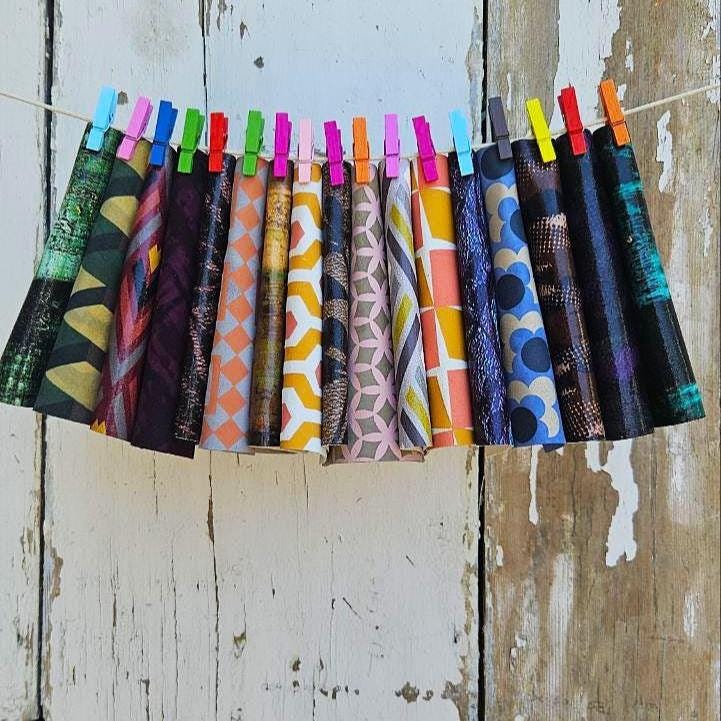 Può includere: Una collezione di coni di tessuto fantasia, ciascuno fissato a una corda con una molletta colorata. I tessuti presentano una varietà di disegni, tra cui forme geometriche e motivi floreali, in una gamma di colori vivaci.