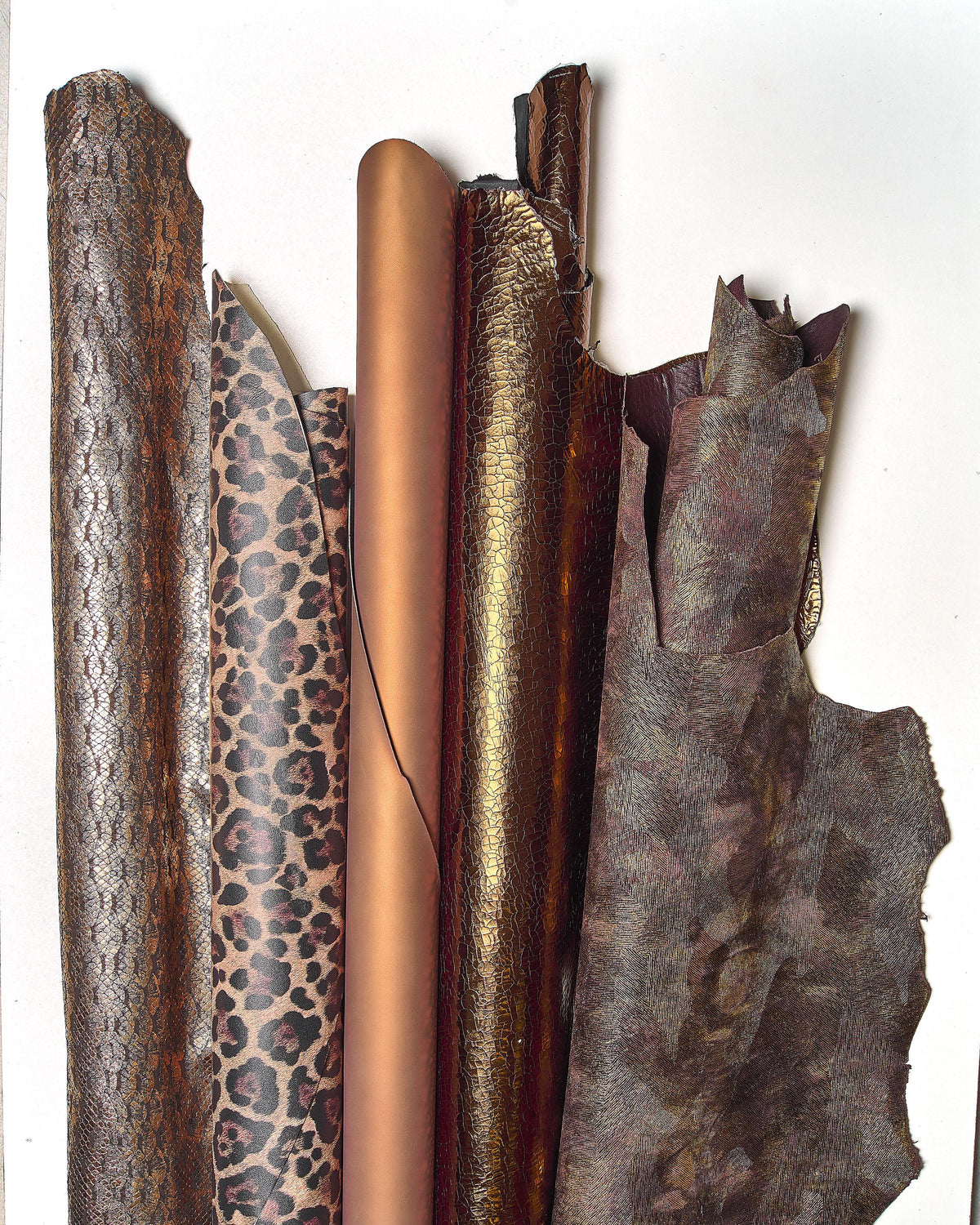 Può includere: Una collezione di pelli di cuoio arrotolate e piatte in vari modelli e colori. Le pelli includono texture a pelle di serpente, stampa leopardata e tonalità bronzo e marrone tinta unita. Le trame variano da lisce a goffrate, mostrando la diversità dei materiali in pelle.