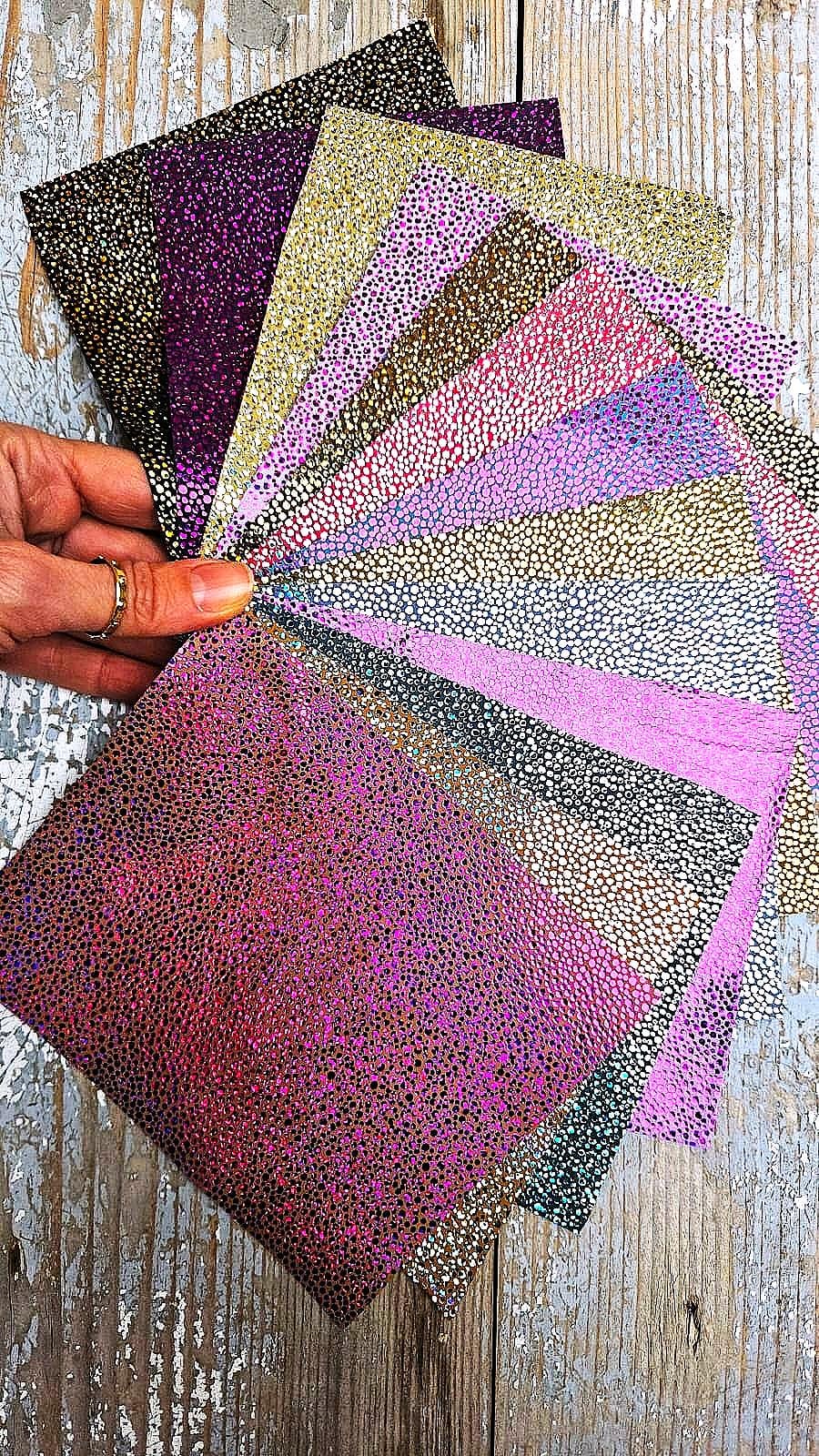 Può includere: Una collezione di carte artigianali colorate e strutturate in tonalità viola, rosa, oro e nero. Le carte hanno un motivo maculato e sono a ventaglio, mostrando la varietà di colori e trame. Le carte sono tenute in mano.