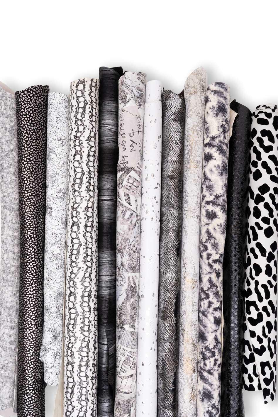 Può includere: Una collezione di undici rotoli di tessuto verticali con vari motivi monocromatici, tra cui stampa leopardata in bianco e nero, texture in pelle di serpente, marmorizzazione grigia astratta, disegni finemente maculati e un rotolo bianco con testo nero.