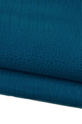 May include: Close-up of teal-colored leather with a crocodile skin texture. The leather is folded, revealing the detailed pattern of scales and ridges. The color is a deep, saturated teal, with subtle variations in tone.