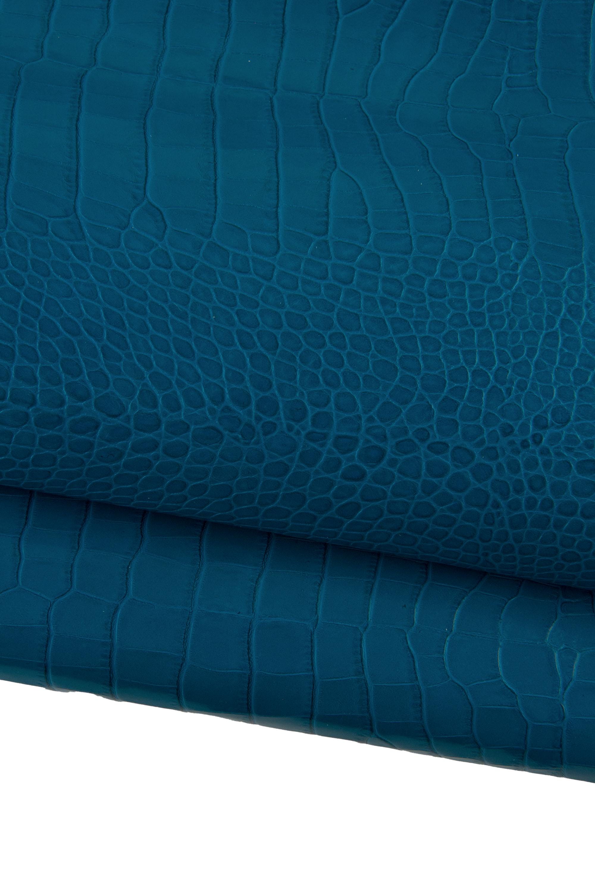 May include: Close-up of teal-colored leather with a crocodile skin texture. The leather is folded, revealing the detailed pattern of scales and ridges. The color is a deep, saturated teal, with subtle variations in tone.