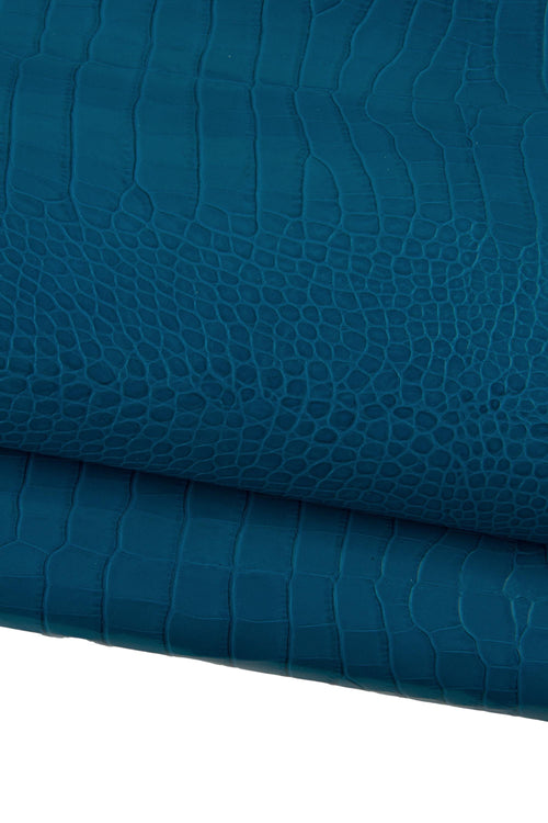 May include: Close-up of teal-colored leather with a crocodile skin texture. The leather is folded, revealing the detailed pattern of scales and ridges. The color is a deep, saturated teal, with subtle variations in tone.