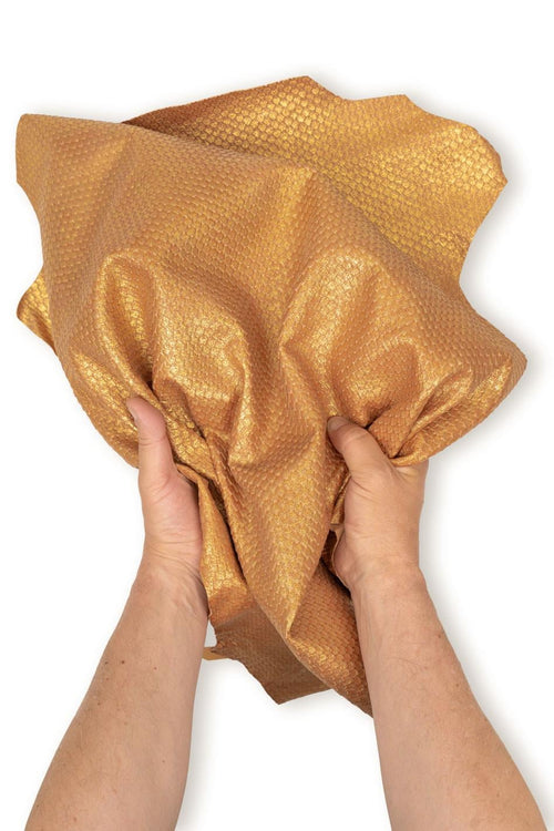 Può includere: Un pezzo di pelle flessibile color oro metallizzato caratterizzato da un distinto motivo a scaglie in rilievo che si ripete su tutta la superficie. Il materiale è mostrato piegato e raccolto, evidenziandone la consistenza flessibile e la finitura lucida.