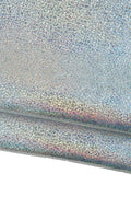 Può includere: Primo piano di un tessuto cangiante e iridescente con una superficie strutturata. Il materiale sembra essere piegato, rivelando strati di argento, blu e accenni di rosa e viola. Il tessuto ha un aspetto maculato, simile a glitter.