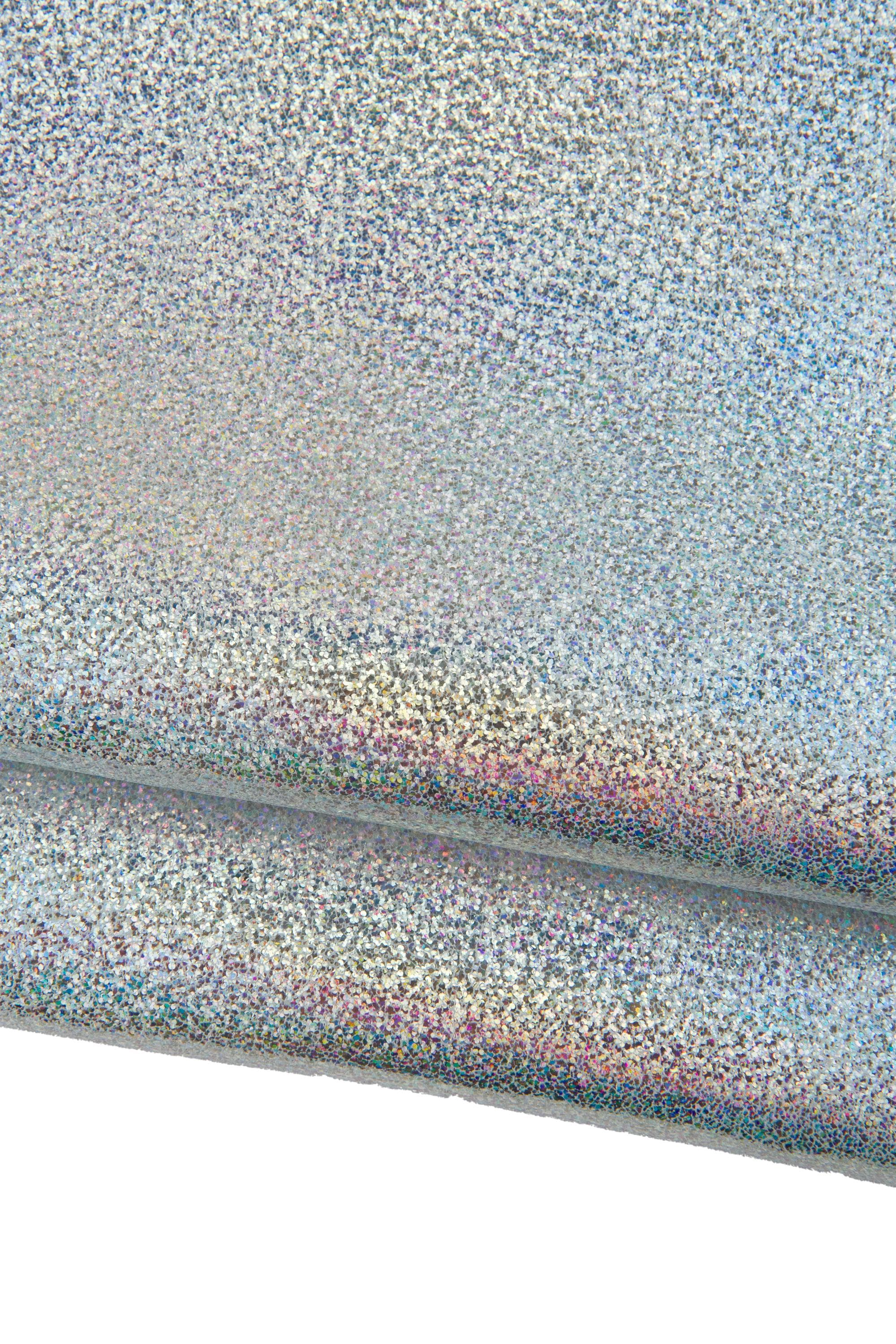Può includere: Primo piano di un tessuto cangiante e iridescente con una superficie strutturata. Il materiale sembra essere piegato, rivelando strati di argento, blu e accenni di rosa e viola. Il tessuto ha un aspetto maculato, simile a glitter.