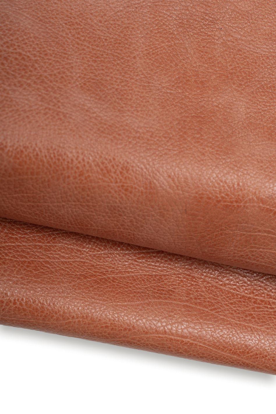 Può includere: Una vista ravvicinata di un pezzo di pelle marrone medio con una texture a grana naturale visibile. Il materiale è ripiegato su se stesso, creando una curva morbida e mostrando la finitura opaca e il motivo superficiale organico della pelle.