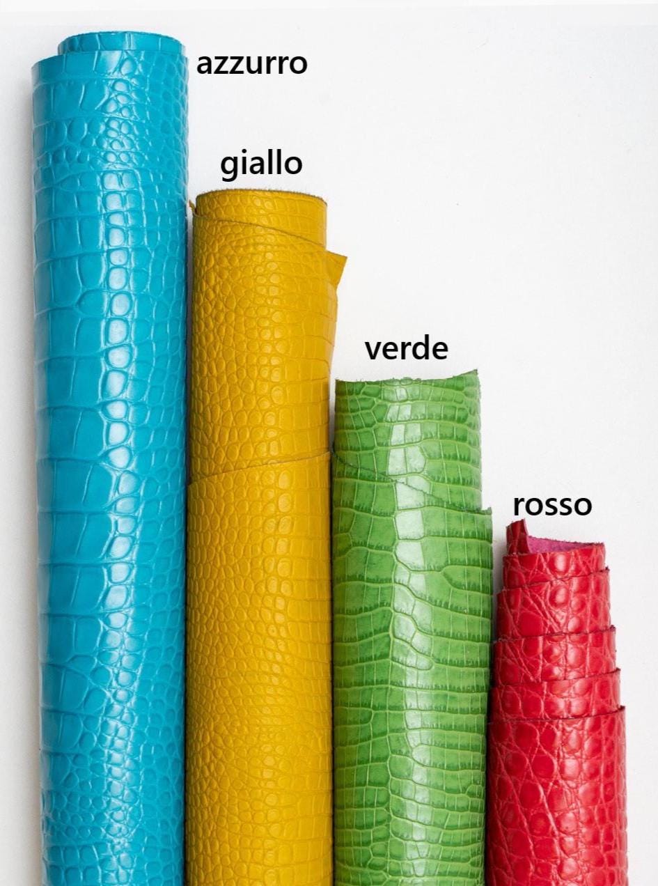 May include: Four rolls of textured leather in vibrant colors: azure, yellow, green, and red. The leather has a crocodile-like pattern. The word "azzurro" is written above the azure roll, "giallo" above the yellow, "verde" above the green, and "rosso" above the red.