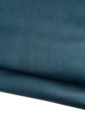 Può includere: Primo piano di un pezzo di pelle piegata color teal. La pelle ha una finitura liscia e opaca e una leggera lucentezza. Le pieghe creano ombre e profondità, evidenziando la consistenza e le variazioni di colore.