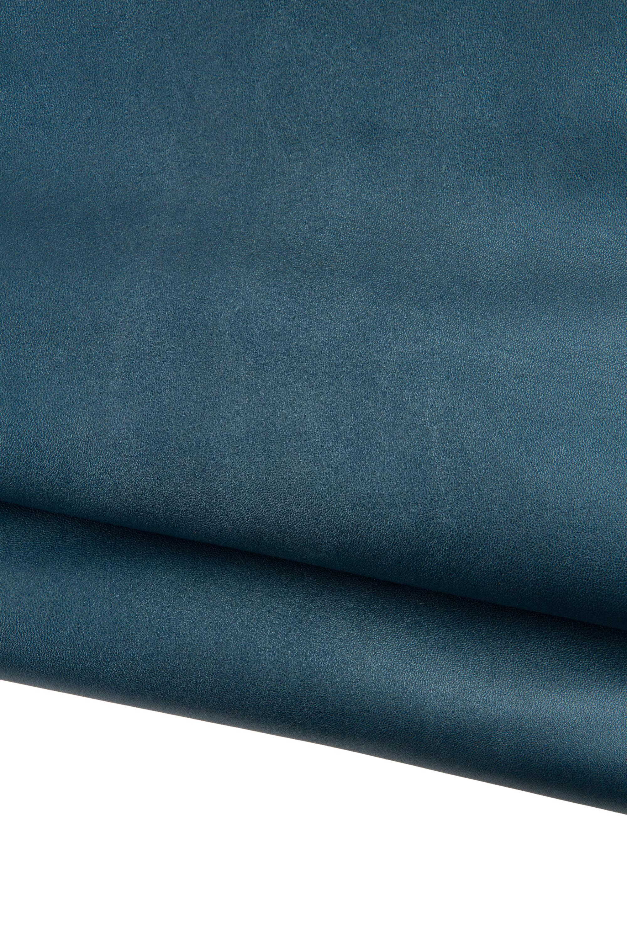 Può includere: Primo piano di un pezzo di pelle piegata color teal. La pelle ha una finitura liscia e opaca e una leggera lucentezza. Le pieghe creano ombre e profondità, evidenziando la consistenza e le variazioni di colore.